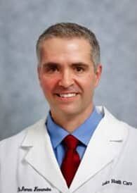 Dr. James Fernandes, DC Headshot photo of Dr. James Fernandes, chiropractor at Eustis Injury Medical Center: Lake Health Care Center, wearing a dress shirt, tie and medical jacket with his name embroidered on the right chest panel and "Lake Health Care Center" on the left chest panel.