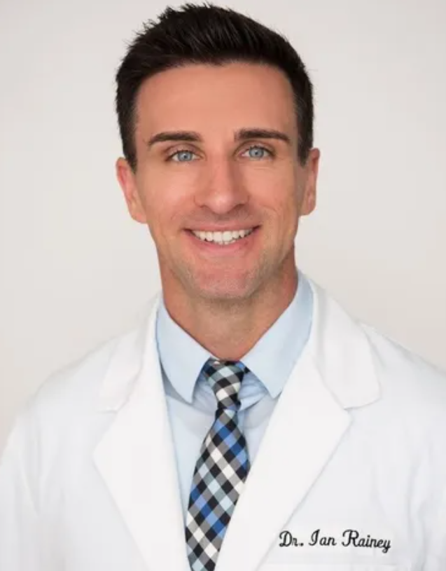Headshot photo of Dr. Ian Rainey, chiropractor at St. Petersburg Medical Injury Center: Rainey Chiropractic. Doctor Rainey smiling and wearing a lab coat with his name embroidered on left side.