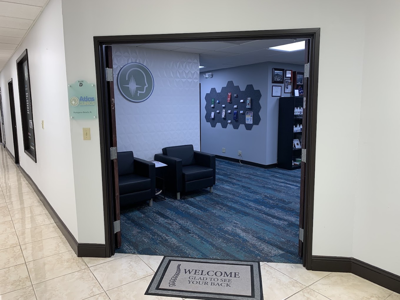 View of interior of Atlas Clinic waiting area from outside the office entrance doors. Double doors to office are open, welcome mat outside doorway, photo taken from the building hallway outside the office entrance