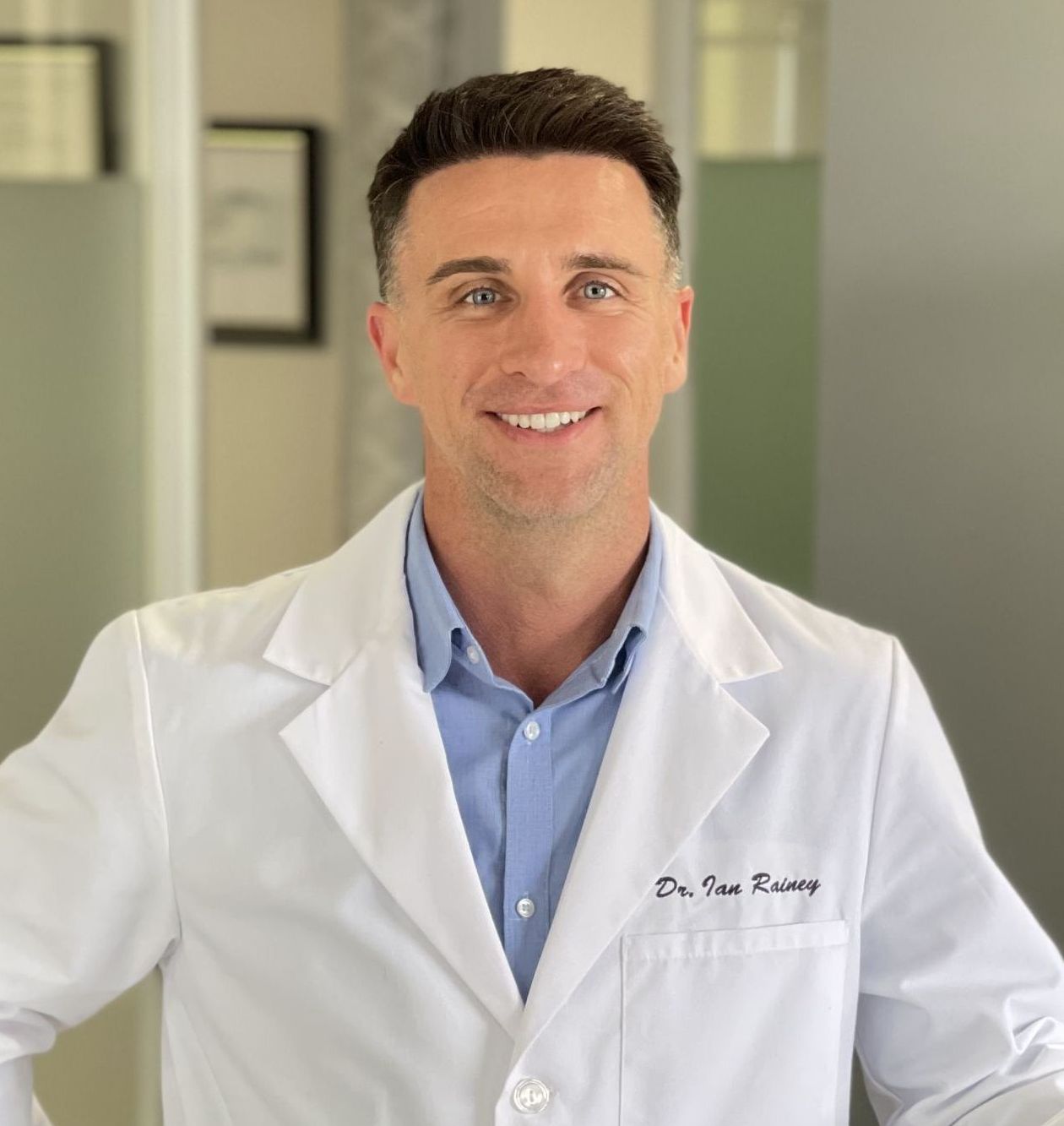 Dr. Rainey of Rainey Chiropractic, smiling and standing with left arm resting on a half wall with right hand on hip; wearing blue shirt and buttoned up medical jacket.