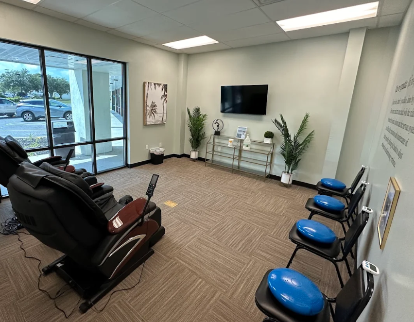 Therapy room with chairs with wobble cushions lined up against one wall, glass wall on opposite side of room, a couple of massage chairs on another side, and a tv on the opposing wall at Panama City Medical Injury Center: Balanced Chiropractic